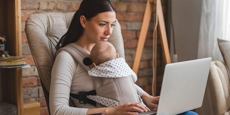 Woman using a laptop while holding a baby in a sling in a cozy living room.